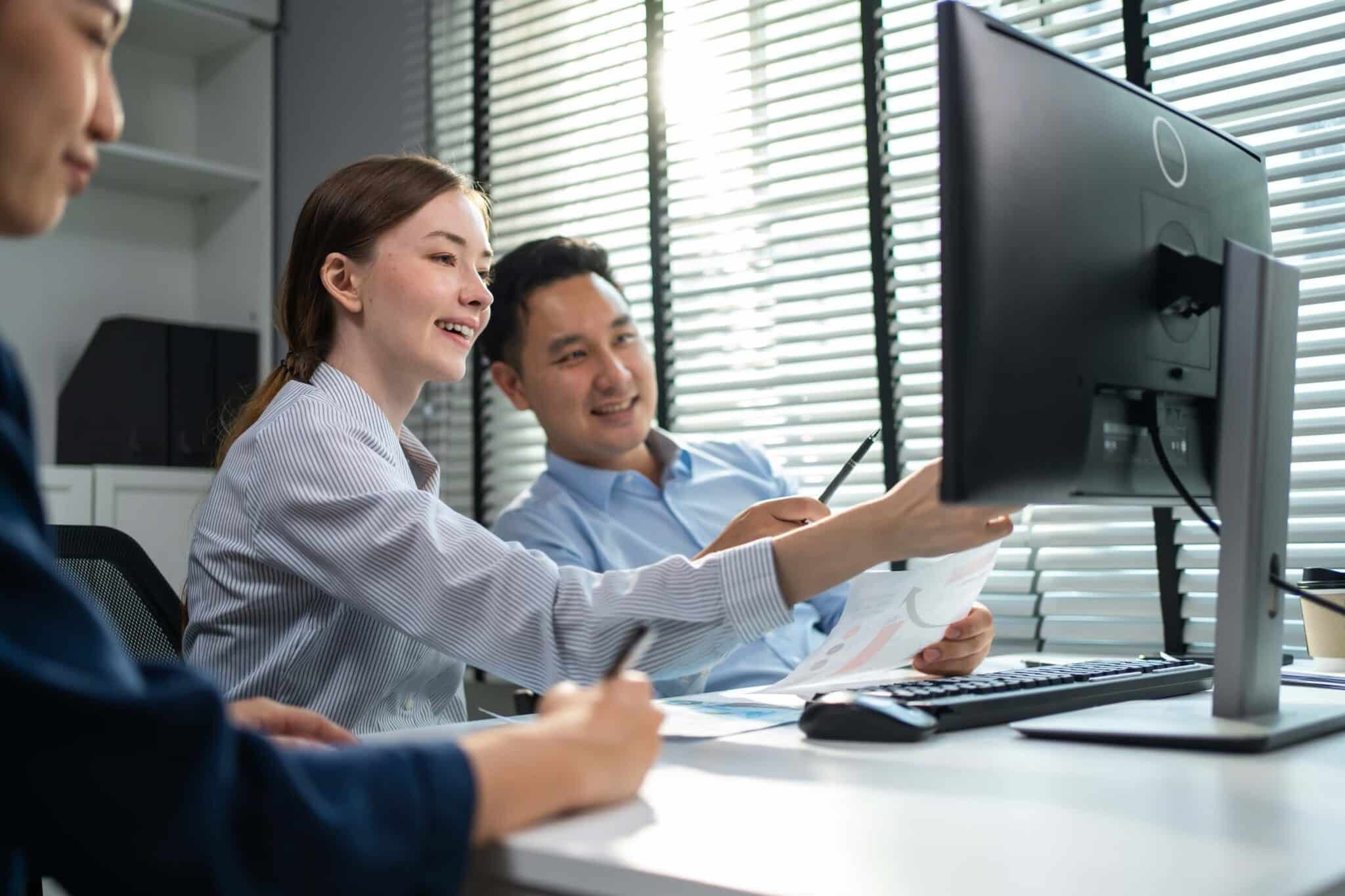 Three colleagues sit at a desk in an office, smiling and looking at a computer monitor. One woman points at the screen while holding papers, engaging the group in discussion about invoice automation as sunlight streams through window blinds behind them.