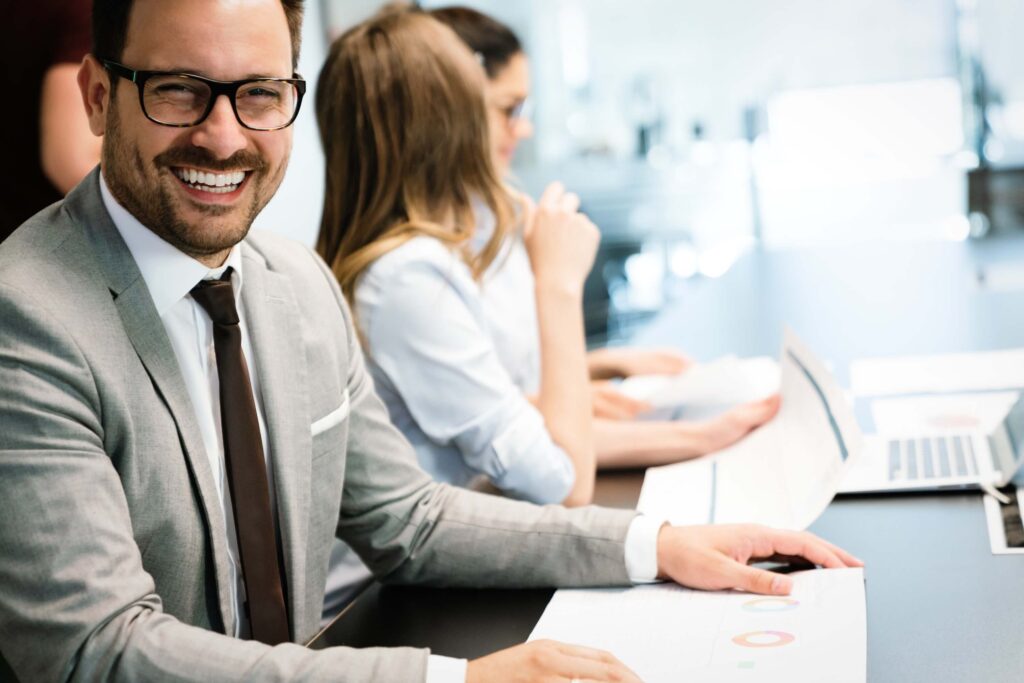 A smiling man in a suit and glasses sits at a desk with papers, while two colleagues work beside him with laptops and documents in a bright, modern office setting.
