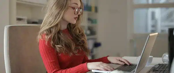 A woman with long blonde hair and glasses, wearing a red sweater, sits at a desk using a laptop in a bright, modern office setting.
