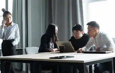 Four people are in a modern office. Three are sitting at a table focused on a laptop, discussing something, while one person stands nearby, talking on a phone. Light streams in from a large window.