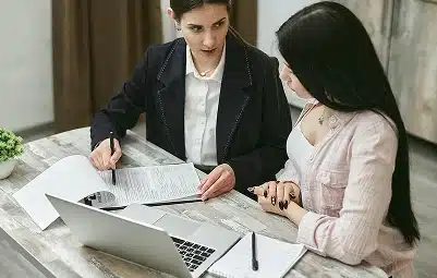 Two women sit at a table with documents, a laptop, and a notepad. One woman, dressed in a blazer, appears to be explaining or reviewing paperwork with the other, who is listening attentively.