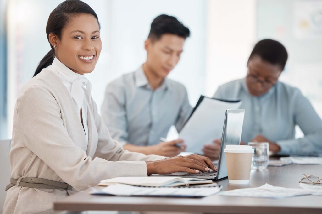 A smiling woman types on a laptop at a desk with documents and a coffee cup, while two colleagues work in the background, reading papers.