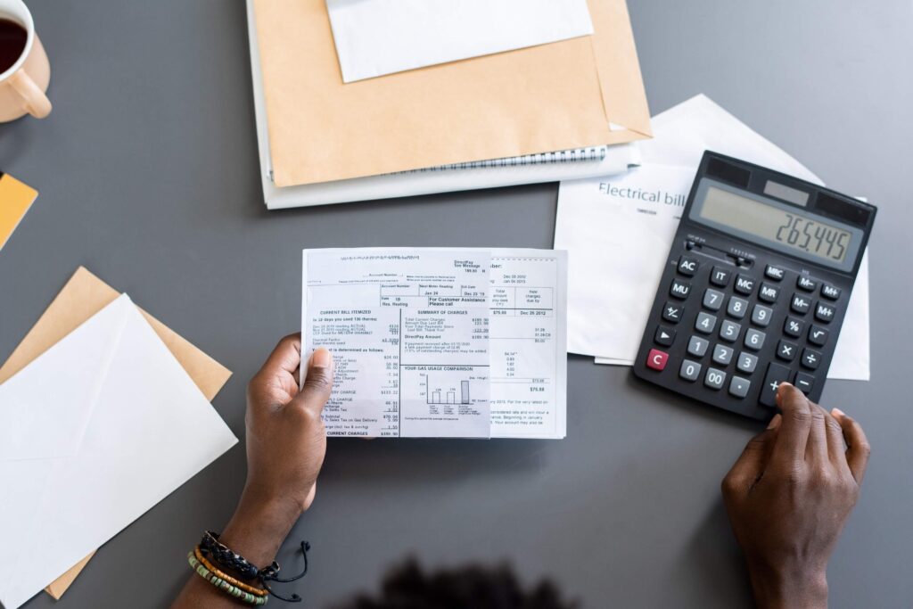 A person holding a utility bill in one hand and using a calculator with the other, surrounded by envelopes, papers, and a cup of coffee on a gray desk.