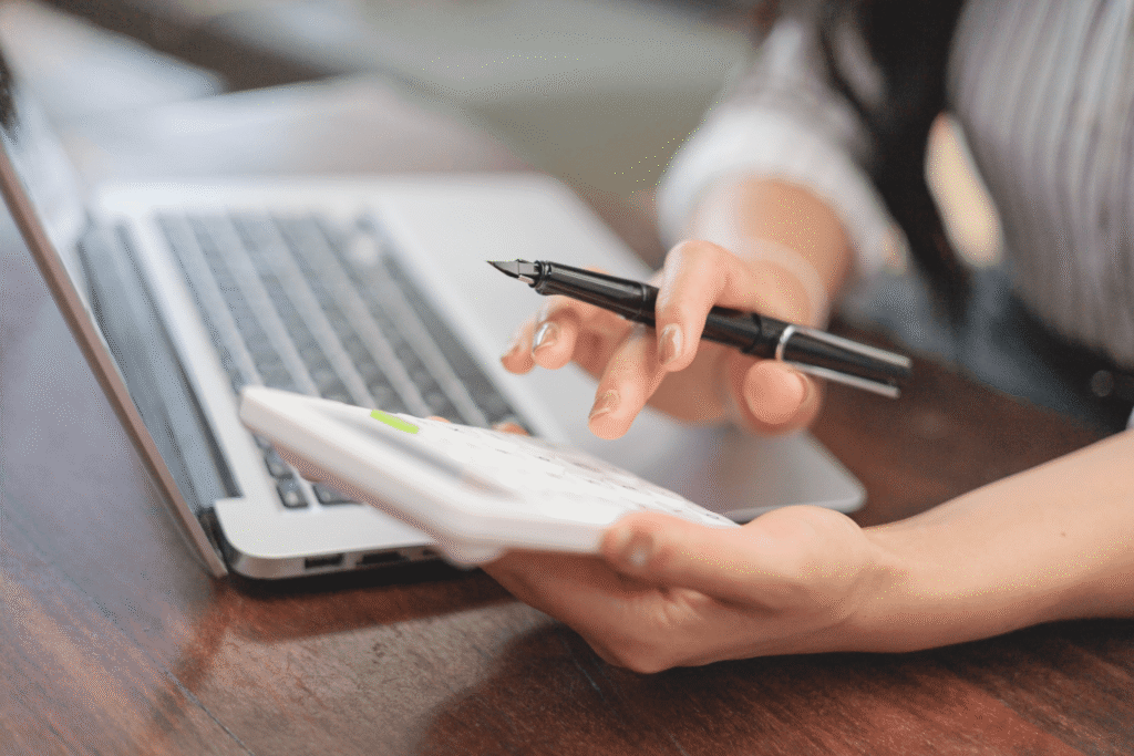 A person holding a pen and using a calculator while working on a laptop at a wooden desk. Only their hands and part of the laptop are visible.