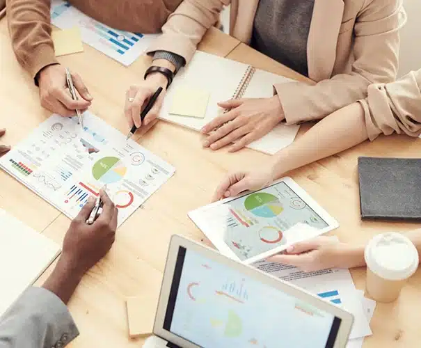 Four people sit around a wooden table discussing charts and graphs. Laptops, papers, notebooks, a gray folder, and a coffee cup are on the table. Only their hands and arms are visible, suggesting a business meeting or teamwork.