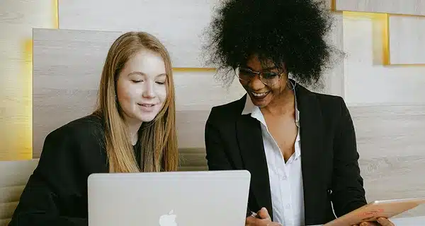 Two women in business attire sit together, smiling while looking at a laptop. One holds a tablet. They appear to be collaborating or discussing work in a modern office setting with light wood paneling.