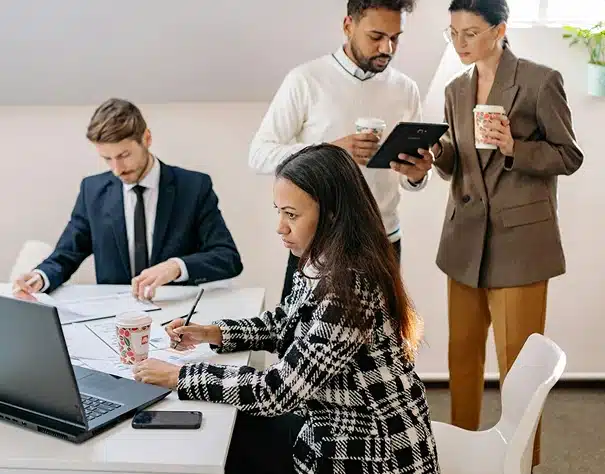 Four people are working in an office. Two are sitting at a table with a laptop and papers, while two others stand nearby with coffee cups, discussing something on a tablet. Everyone appears focused and engaged.