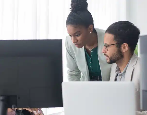 Two people in business attire work together at a desk, looking intently at a computer monitor in a bright office setting. One person is seated while the other is standing and leaning in to observe the screen.