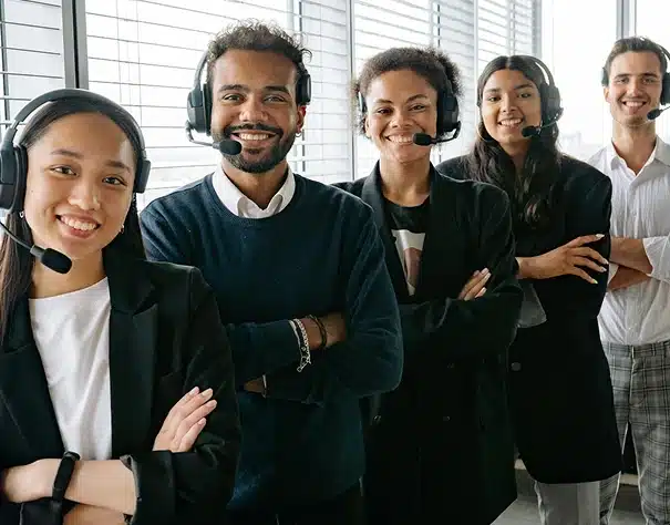 Five customer service representatives wearing headsets stand in a row, smiling confidently with arms crossed, in a modern office with large windows and blinds.