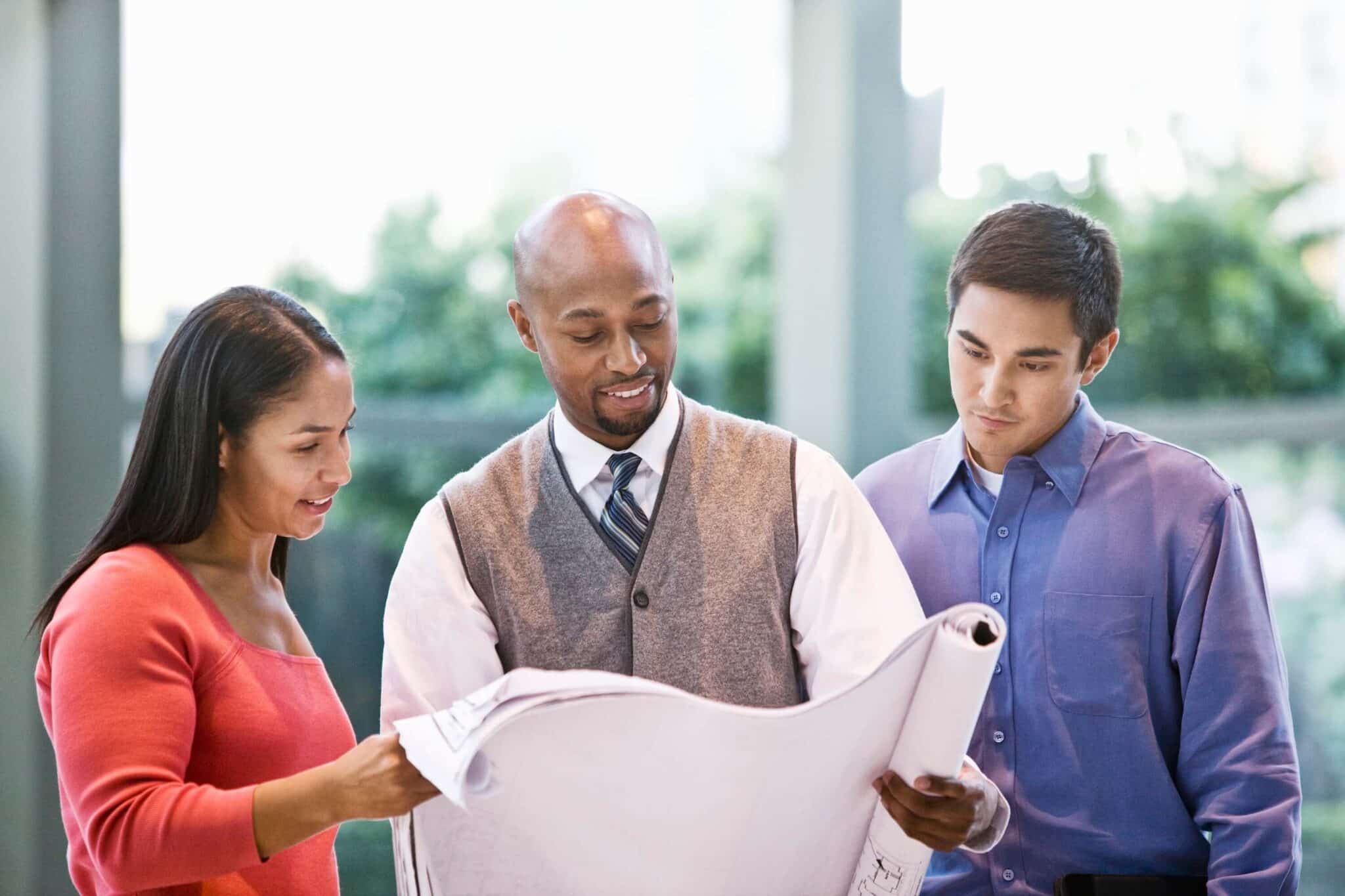 Three people, two men and one woman, stand together indoors, focused on large architectural blueprints. They appear engaged in discussion about project cost overrun, with large windows and greenery visible in the background.