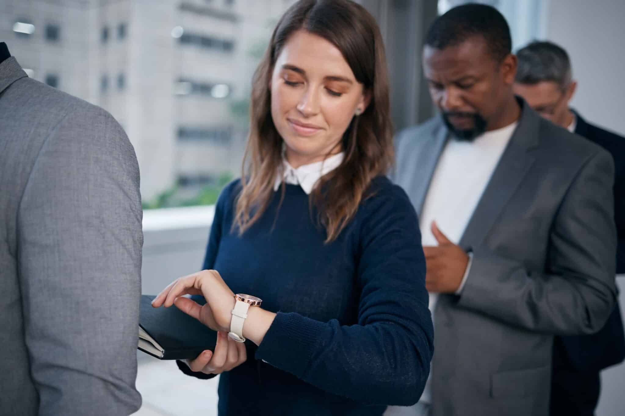 A woman standing in line looks at her watch while holding a notebook—perhaps pondering how to get employees to submit timesheets on time—with two men behind her also waiting inside a modern office building.