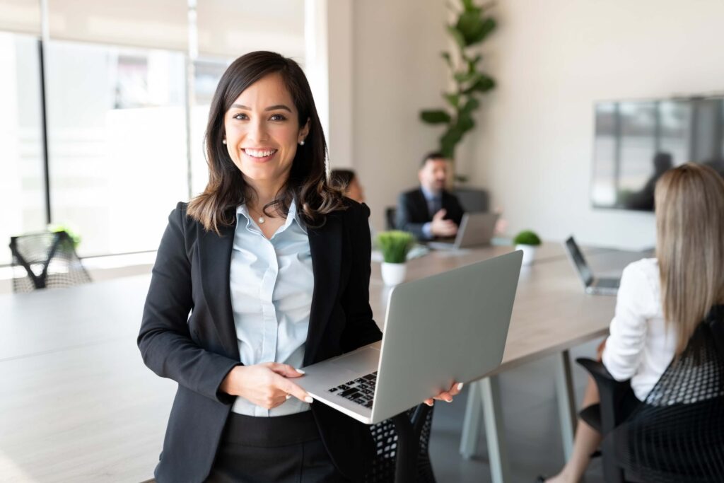 A smiling woman in business attire holds a laptop in a modern office, with colleagues working at a conference table in the background.