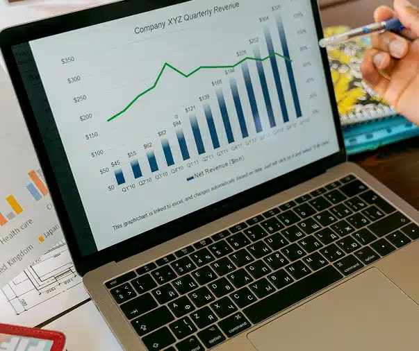 A laptop displays a chart titled "Company XYZ Quarterly Revenue" showing a bar and line graph with data from Q1'19 to Q3'21. A person holding a pen points at the screen. Papers and a notebook are nearby.