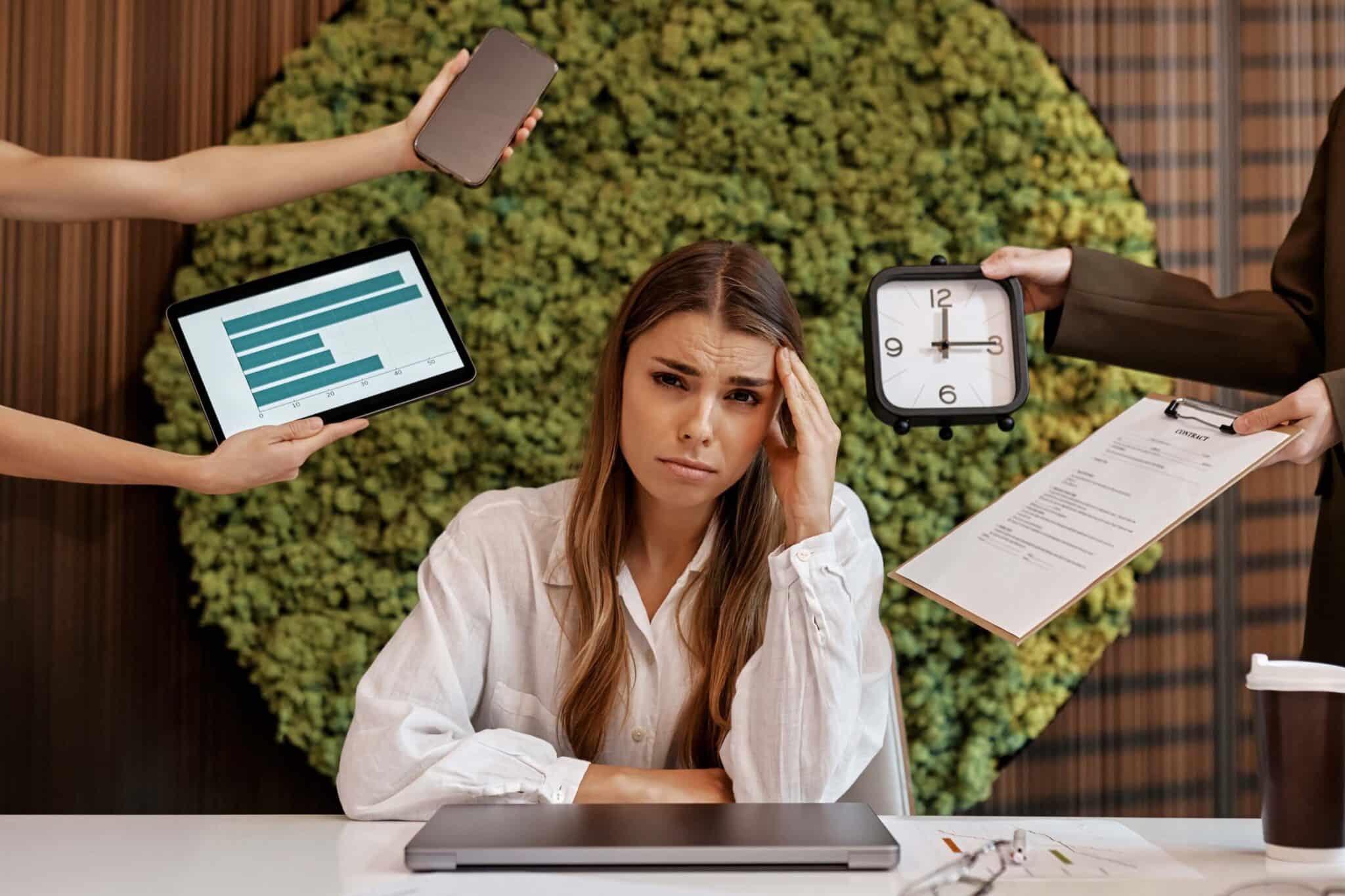 A stressed woman sits at a desk with her hand on her forehead as people around her hold out a phone, a tablet with a graph, a clock, and a clipboard—one showing a timesheet error—highlighting how she's overwhelmed by multiple tasks.