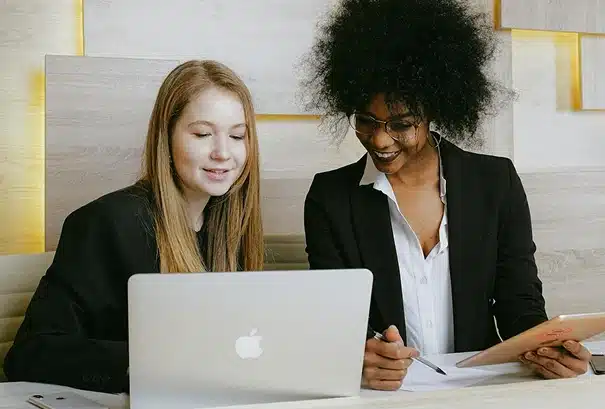 Two women in business attire sit at a desk; one uses a laptop while the other writes on a notepad. They appear to be collaborating or discussing something on the laptop screen (enterprise time and attendance software).
