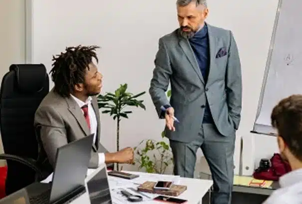 Two men in suits have a serious discussion in an office setting, with one standing and gesturing toward the other, who is seated at a desk with laptops and documents. A flip chart and plant are visible in the background.