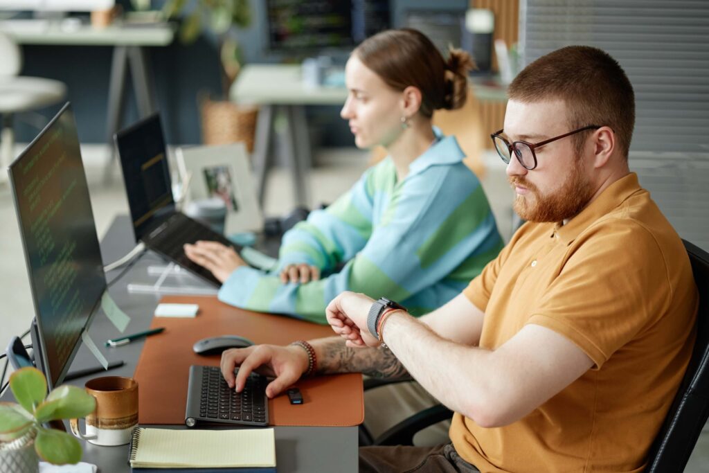 A man in glasses checks his smartwatch, possibly tracking the pay period, while typing at his desk. Beside him, a woman works on a laptop. Computer screens, notebooks, and plants are visible on their desks as they prepare to submit timesheets on time.