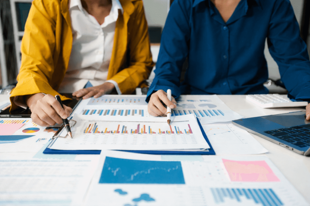 Two people review charts and graphs on paper at a desk, analyzing financial data with pens in hand. A laptop, keyboard, and colorful documents suggest a focus on finance workflow automation to streamline financial processes.
