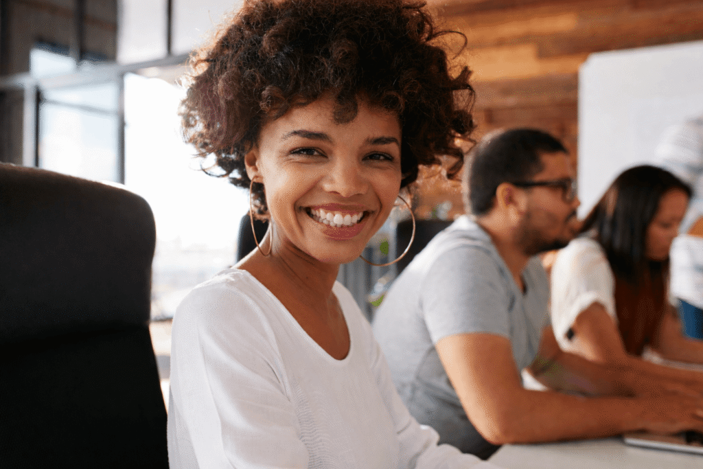 A woman with curly hair and hoop earrings smiles at the camera while sitting at a desk in a modern office, ready to tackle expense reports. Two coworkers are in the background, working on their computers.