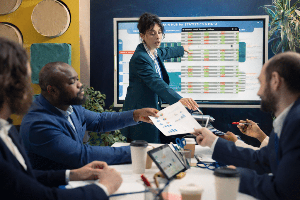 A businesswoman in a suit points to a digital screen showing charts and data on financial reporting while four colleagues sit around a table with documents, tablets, and coffee cups, engaged in a discussion.