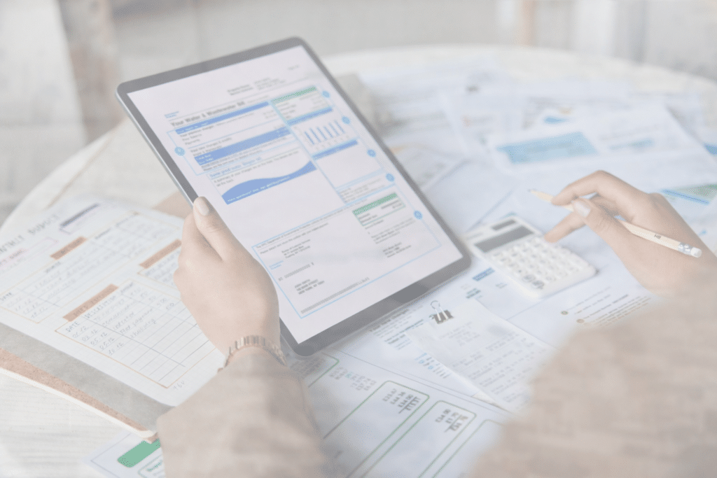 A person uses a tablet and a calculator while reviewing financial documents and charts spread out on a table. The scene suggests working on budgeting, accounting, or financial planning tasks.