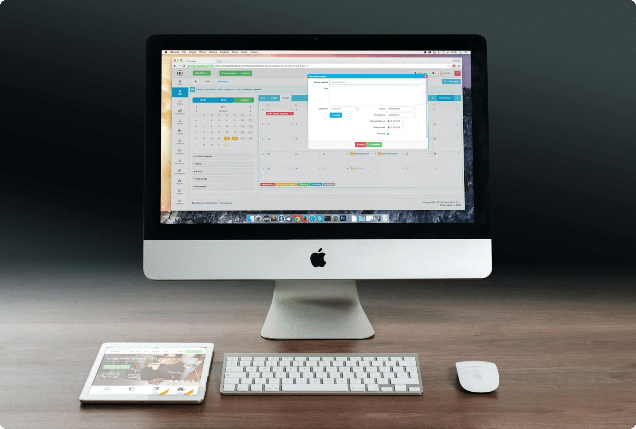 A desktop computer displaying a calendar and scheduling application is on a wooden desk, alongside a wireless keyboard, mouse, and a tablet showing a website with images and text.