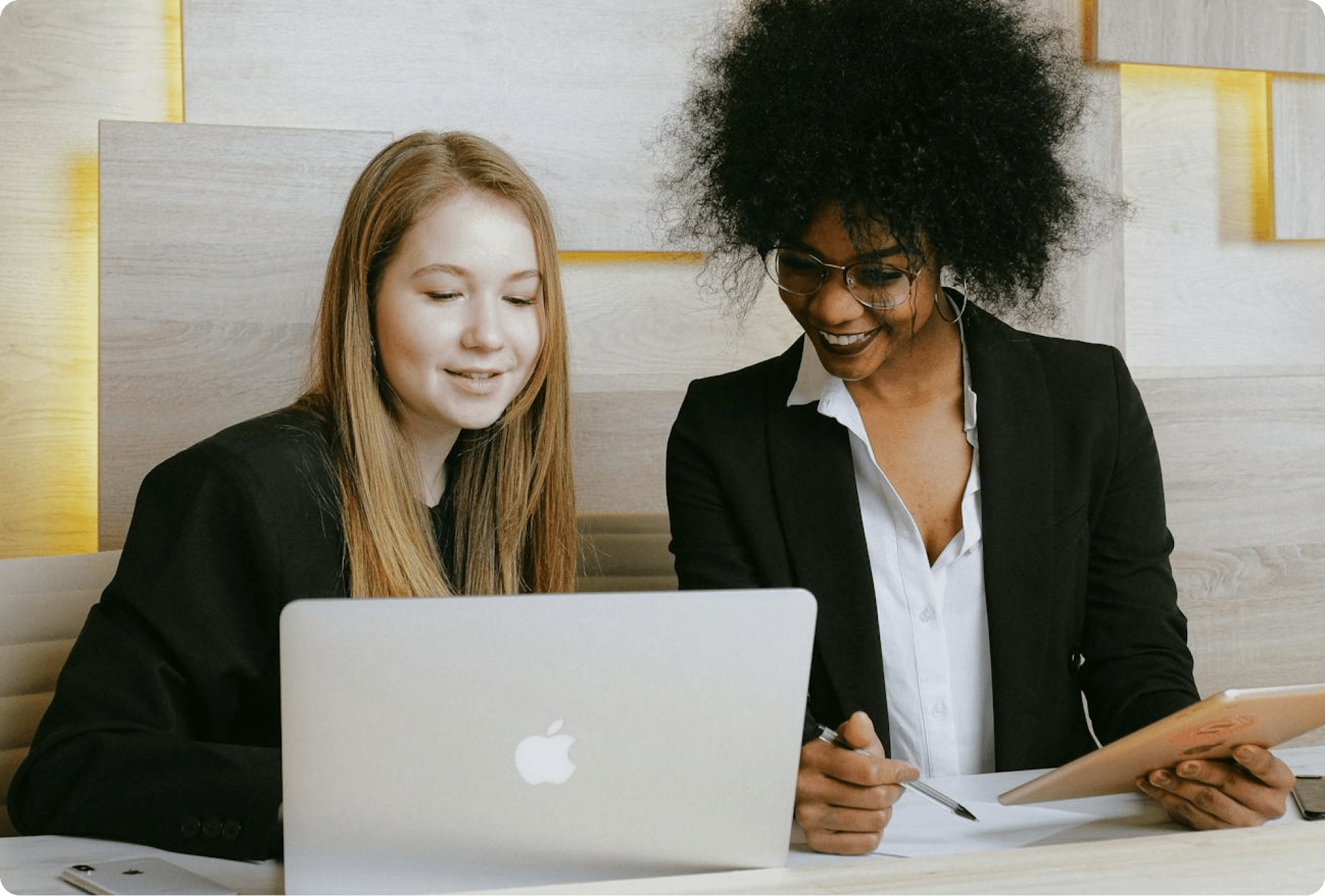 Two women in business attire sit together at a desk; one is using a laptop, while the other holds a tablet and pen. They appear to be collaborating and smiling in a modern office setting.