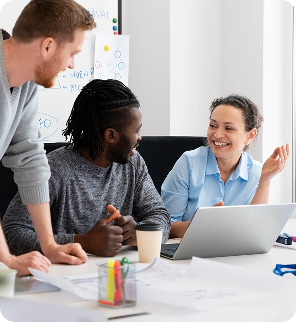 Three colleagues are gathered around a desk with papers and a laptop, smiling and engaging in a lively discussion in a bright, modern office setting.