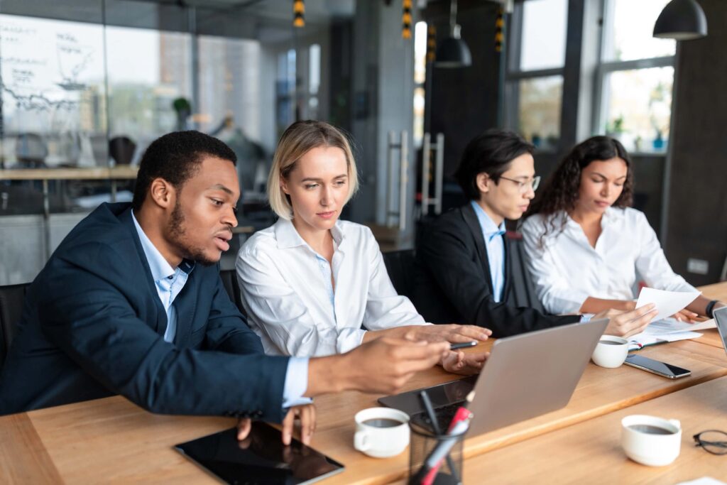 Four business professionals sit at a wooden table with laptops, papers, and coffee cups, discussing the expense approval process. One man points at a laptop screen while the others listen and review expense reports in a modern office setting.