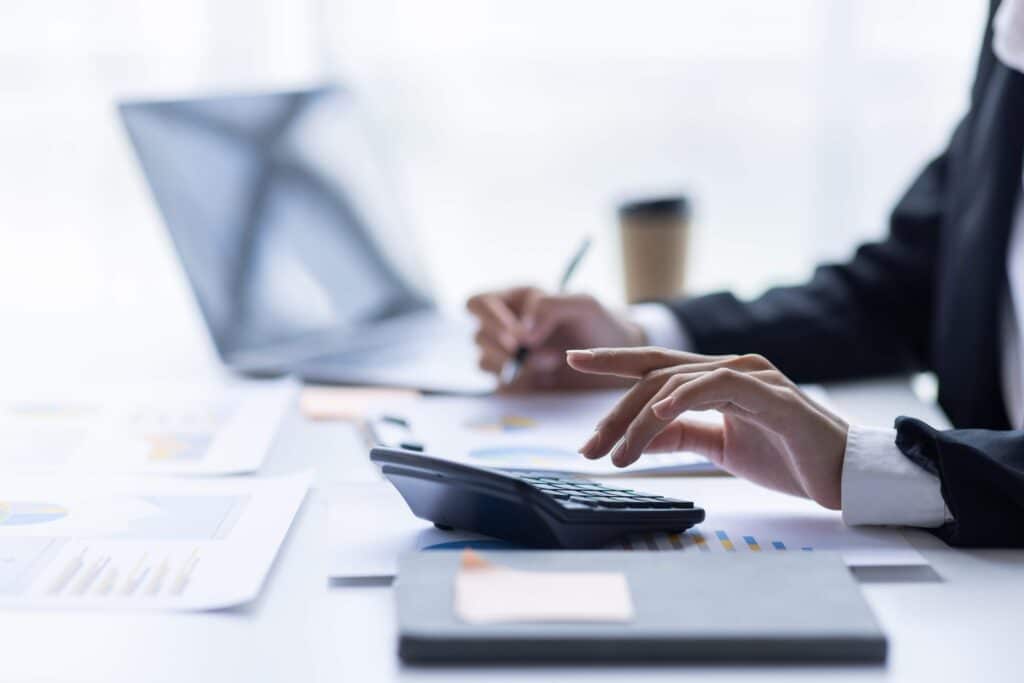 A person in business attire uses a calculator while writing on a document at a desk with papers, a laptop, and a notebook, suggesting finance workflow automation in handling financial processes efficiently.