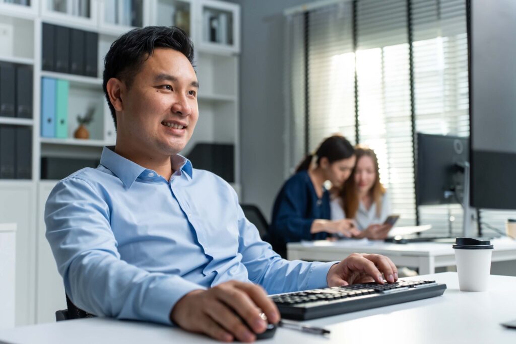 A man in a blue shirt smiles while working at a computer in a bright office. Two women in the background collaborate at a desk, and sunlight filters through window blinds. A coffee cup is on the table.