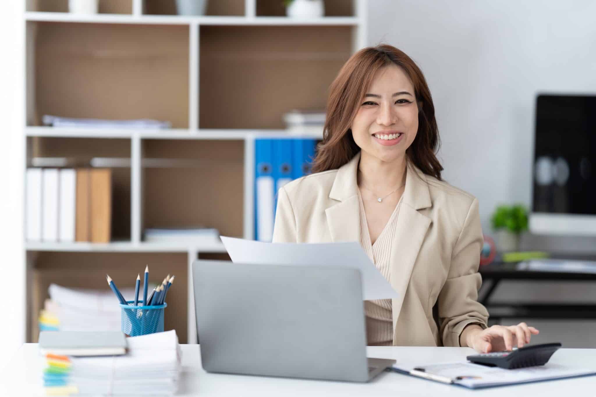 A smiling woman in a beige blazer sits at a desk with a laptop, examining papers and using a calculator—perhaps reviewing an unbalanced balance sheet. Office supplies and shelves are visible in the background.