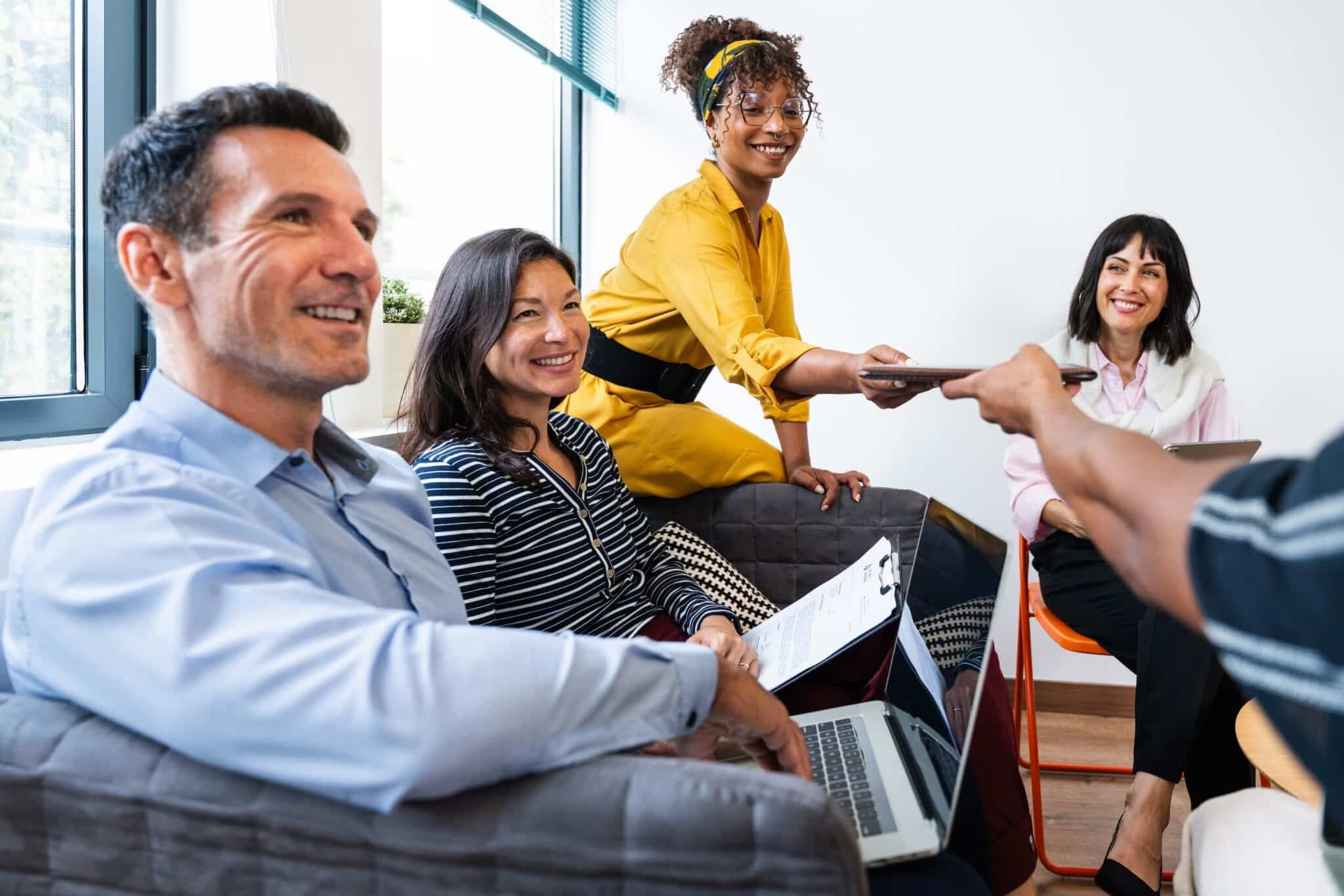 Four people are sitting in a casual meeting room, smiling and talking. One person in a yellow dress stands, passing a tablet for expense approval. Laptops and notepads are visible, reflecting the friendly, collaborative atmosphere.