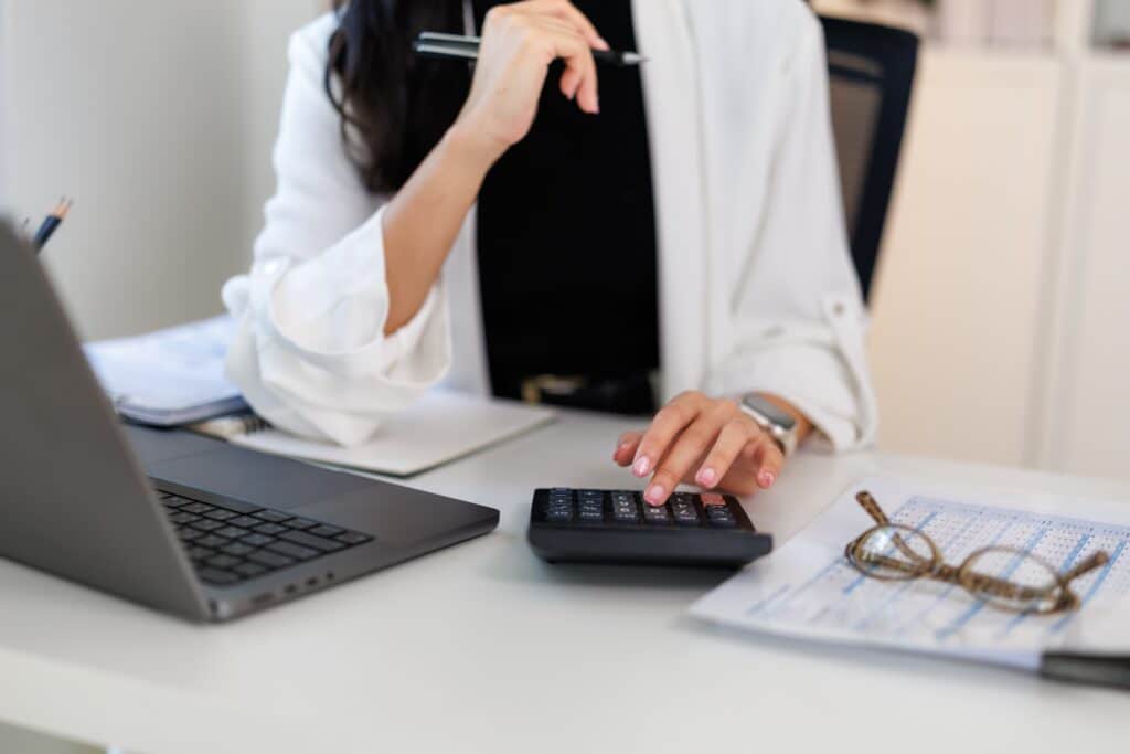 A woman in a white blazer sits at a desk with a laptop, holding a pen and using a calculator. Papers and glasses are on the desk, suggesting she is working on financial tasks.