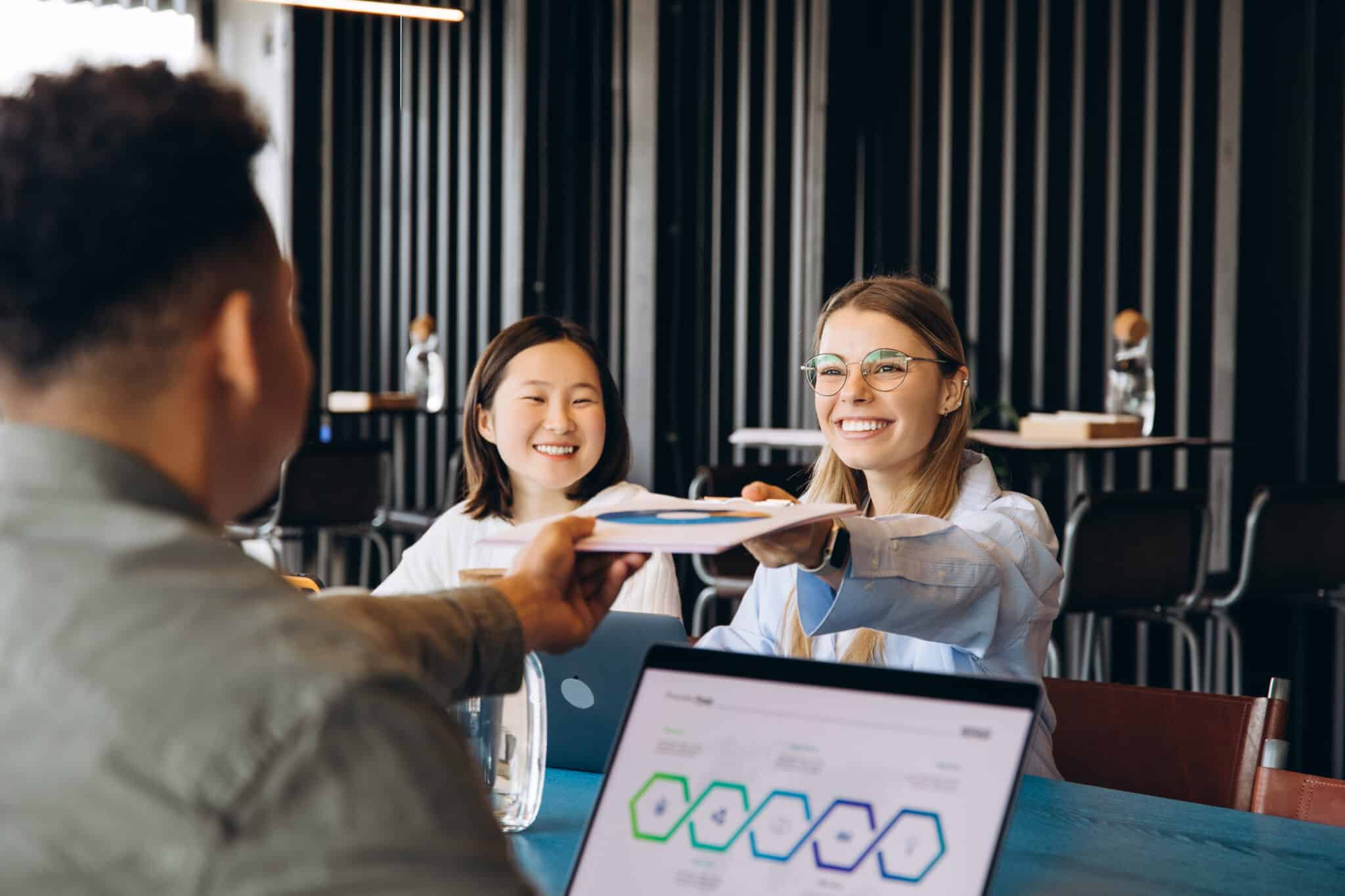 Two women sit at a table in an office, smiling as one hands a document to a man across from them. A laptop displaying a revenue recognition chart is in the foreground, creating a friendly and collaborative atmosphere.