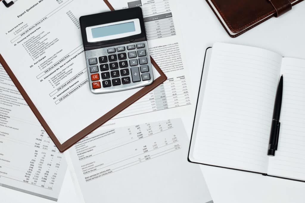 A calculator, financial documents, a brown notebook, and an open blank notebook with a pen are arranged on a white desk, suggesting a workspace for accounting or financial planning.