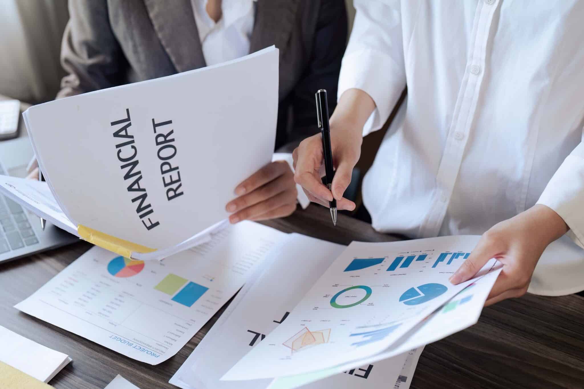 Two people reviewing financial documents at a desk, one holding a “Financial Report” and the other analyzing papers with graphs and charts, carefully checking for financial reporting errors in the financial statements.