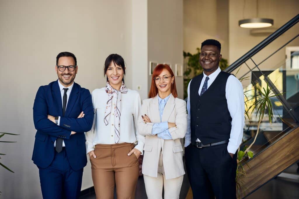 Four professionally dressed people stand side by side in an office, smiling at the camera. The background includes stairs, plants, and modern decor, suggesting a business or corporate environment.