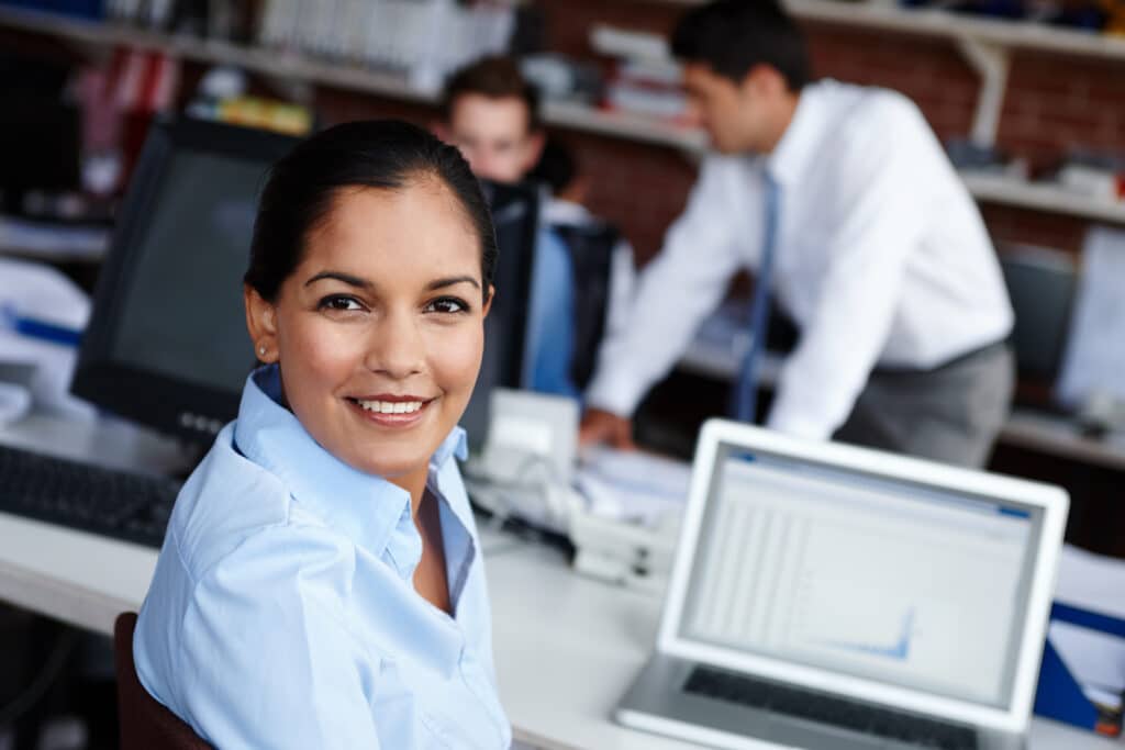 A woman in business attire smiles at the camera while sitting at a desk with a laptop displaying a revenue recognition chart. Two colleagues work in the background in a modern office setting.
