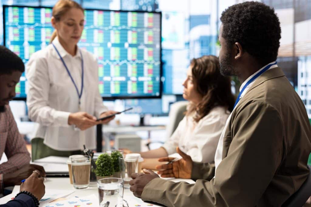 Four people in business attire meet in a modern office. A woman stands holding a tablet as three others sit at the table, discussing finance workflow automation. Stock market data is displayed on large screens in the background.