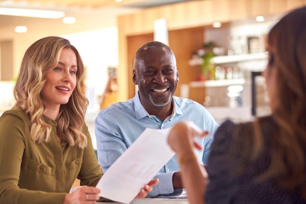 Two people, a woman and a man, sit at a table smiling and looking at a third person holding a document, suggesting a meeting or discussion about the expense approval process in a bright, modern office setting.