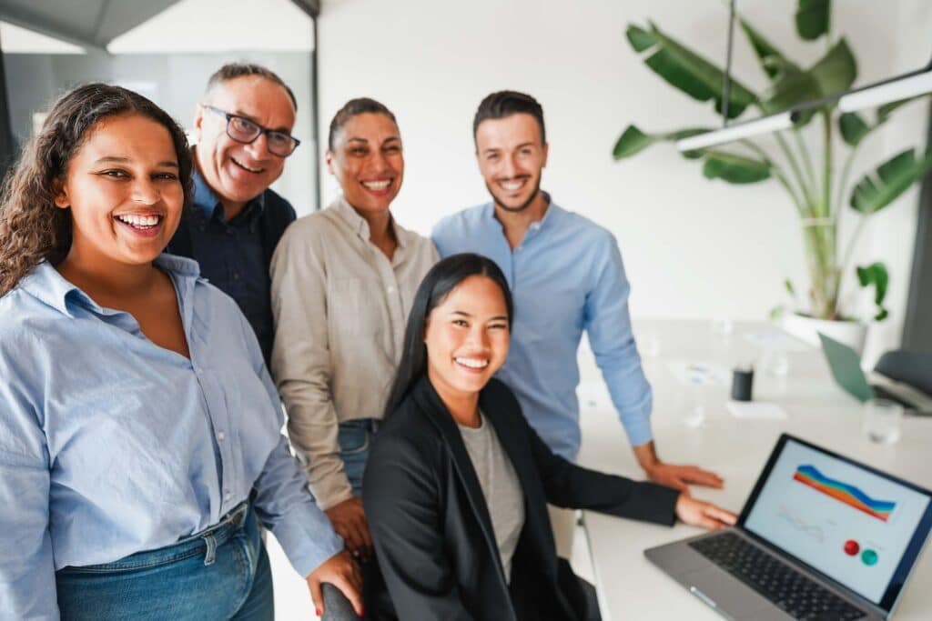 Five people in business attire smile at the camera in a modern office. One person sits at a desk with a laptop displaying colorful graphs, reviewing financial statements. A large green plant and bright windows are in the background.