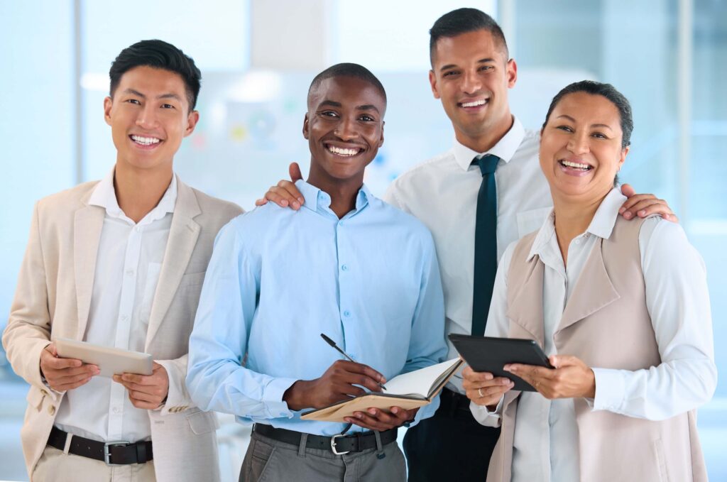 Four professionally dressed people stand together smiling in an office setting. Two hold tablets displaying financial statements, one writes in a notebook, and they appear relaxed and happy, suggesting a collaborative business environment.