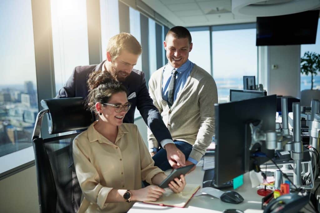 Three colleagues in a bright office gather around a desk, discussing finance workflow automation. One woman sits smiling with a tablet while two men stand beside her, one pointing at the screen. Office equipment and city-view windows complete the scene.