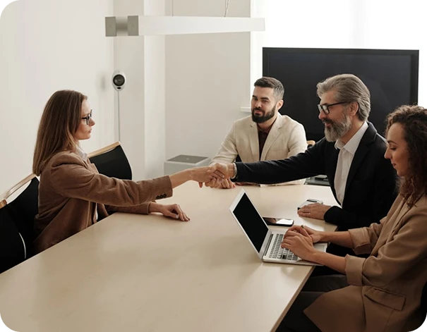 Four people in business attire sit around a conference table. One woman reaches across to shake hands with a bearded man, while two others, a man and a woman with a laptop, look on and smile.