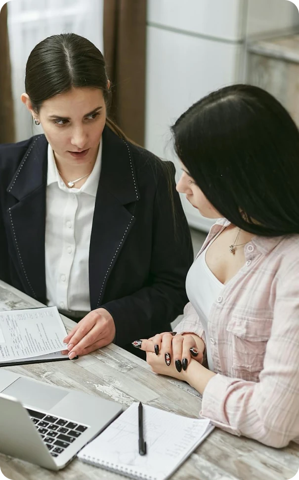 Two women sit at a table with documents and a laptop, engaged in a serious conversation. One woman holds a paper and gestures, while the other listens attentively with her hands folded.