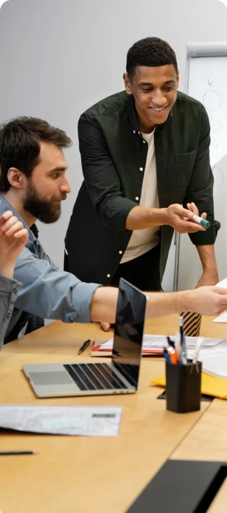 Two men collaborate at a table with a laptop, documents, and office supplies. One man stands and smiles while pointing at a paper, while the other sits and listens attentively. A whiteboard is visible in the background.