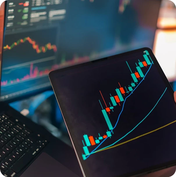 A tablet displays a colorful candlestick chart with trend lines, showing a rising stock price. In the background, a laptop screen also shows financial charts and data.