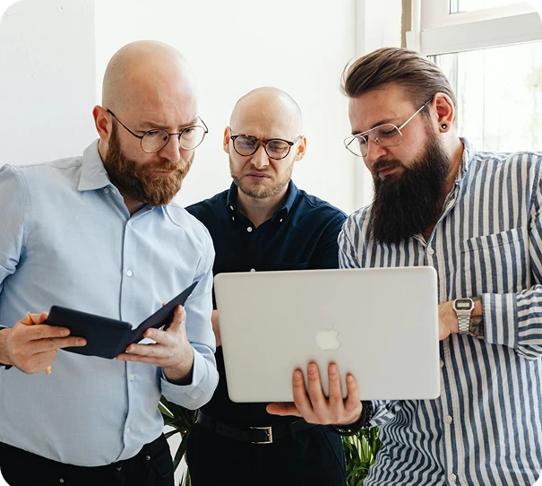 Three men with beards and glasses stand together indoors, looking intently at a laptop screen. One holds a notebook and pen, while another points to the screen. Sunlight comes in through a nearby window.