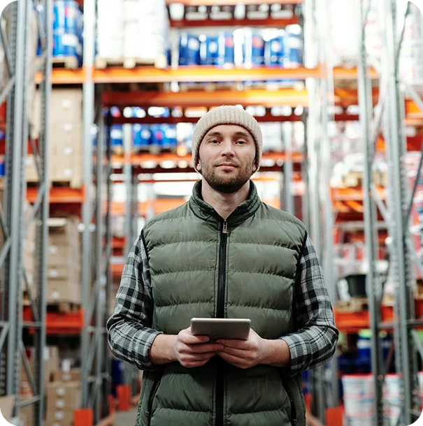 A man wearing a beanie and puffer vest holds a tablet while standing in a warehouse aisle surrounded by tall shelves stacked with boxes and containers.